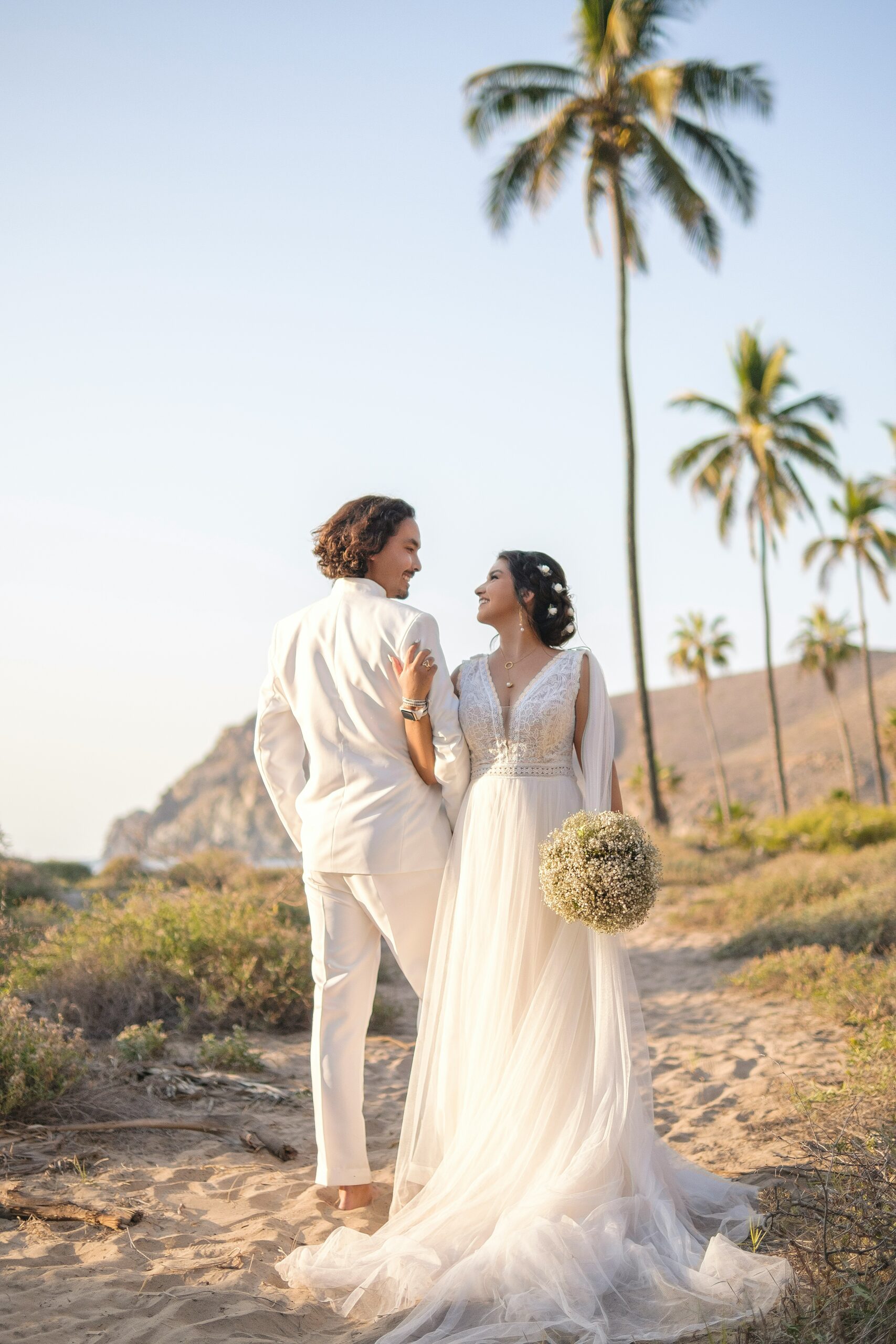 Bride and groom in wedding attire on a sandy beach with palm trees, gazing at each other; bride holds a bouquet in a long tulle gown.
